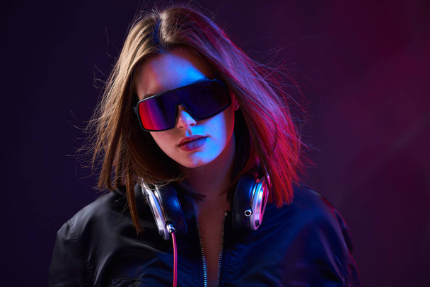 Portrait of stylish DJ girl with headphones, glasses and jacket in nightclub in smoke on a dark background in neon light - Foto, afbeelding