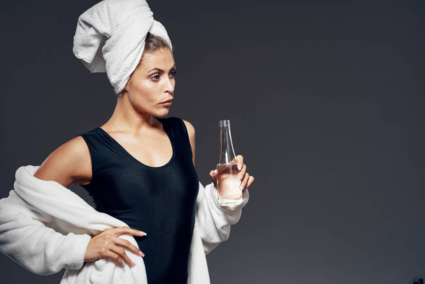 Στιγμιότυπο. Νεαρή γυναίκα σε φόρμα με μπουρνούζι και μπουκάλι νερό σε σκούρο φόντο   - Φωτογραφία, εικόνα
