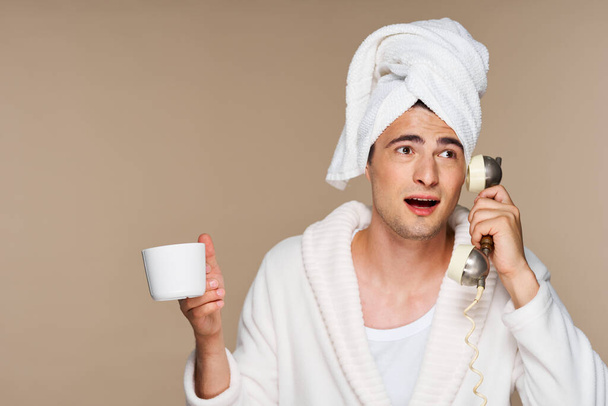 Młody emocjonalny mężczyzna w szlafroku z filiżanką kawy rozmawiający przez zabytkowy telefon. Zdjęcia studyjne. Izolacja - Zdjęcie, obraz