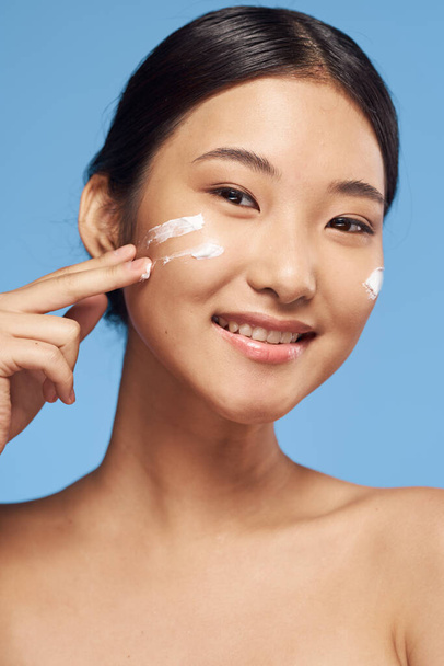 молодая азиатка наносит крем на лицо в студии на синем фоне  - Фото, изображение