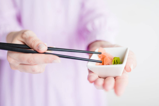 Close up of woman holding bowl with wasabi and gari and chopsticks with sushi roll, japanese traditional healthy meal concept. - Фото, изображение