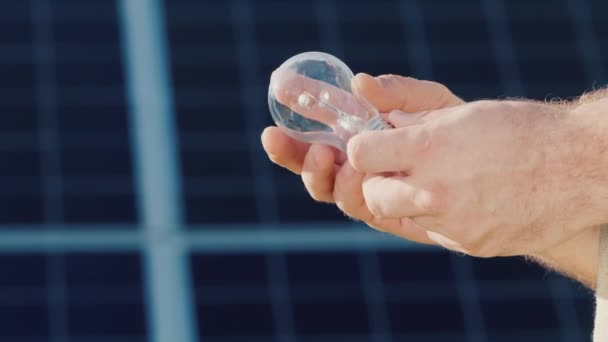 Side view: Mans hands hold a light bulb against the background of solar panels - Felvétel, videó