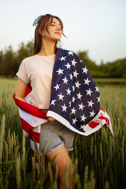 young beautiful caucasian teenager girl in pink top and jeans shorts with national american flag on her in green wheat field at sunset. Independence day - Zdjęcie, obraz