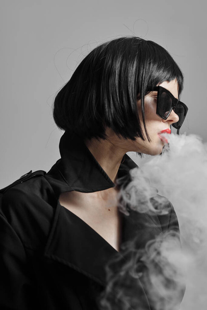 Fiatal, gyönyörű nő napszemüvegben dohányzik a stúdióban. Divatfotózás  - Fotó, kép