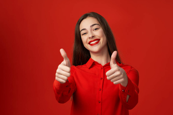 Studio shot. Young beautiful woman in red gesturing thumbs up  isolated on red background - Foto, Bild