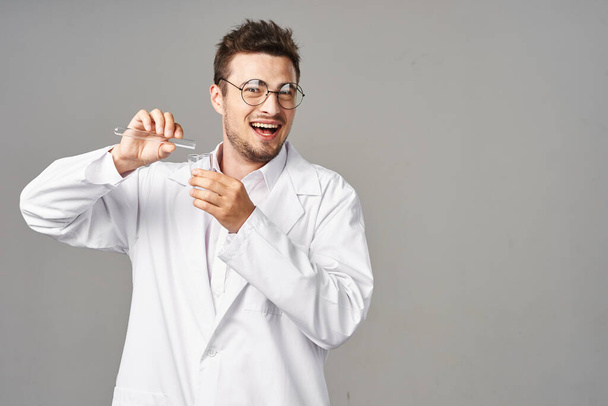 Молодой красивый улыбающийся доктор в халате с пробирками на сером фоне - Фото, изображение