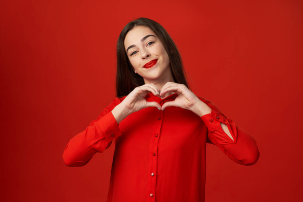 Tiro no estúdio. Jovem mulher bonita em vermelho mostrando coração isolado no fundo vermelho
 - Foto, Imagem
