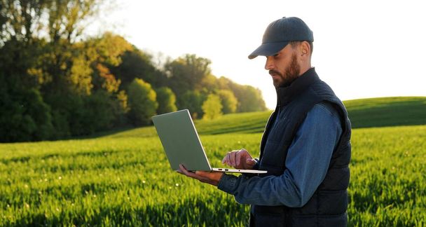 Caucasian young male farmer in hat standing in field and typing on keyboard of laptop computer. Attractive man using device in margin in summer and browsing online. Agricultural concept. - Фото, изображение