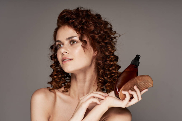 Mujer hermosa joven con cabello rizado sosteniendo spray para el cabello y peine de madera. Captura de estudio
 - Foto, Imagen