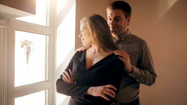 Jovem homem amoroso abraçando sua esposa olhando pela janela ao pôr do sol
 - Foto, Imagem