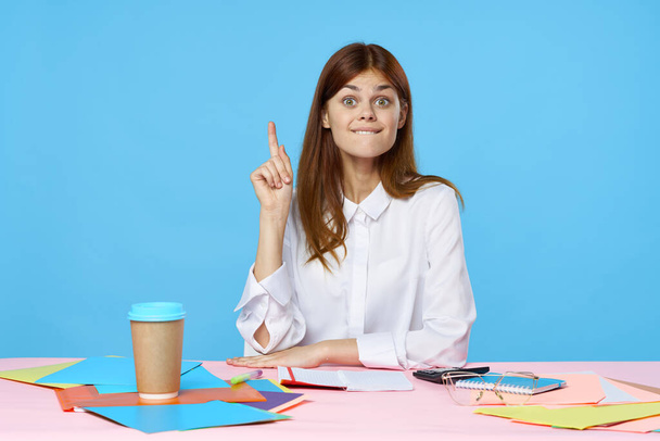 Молода бізнес-леді сидить за столом, вказуючи вгору ізольовано на синьому фоні
  - Фото, зображення