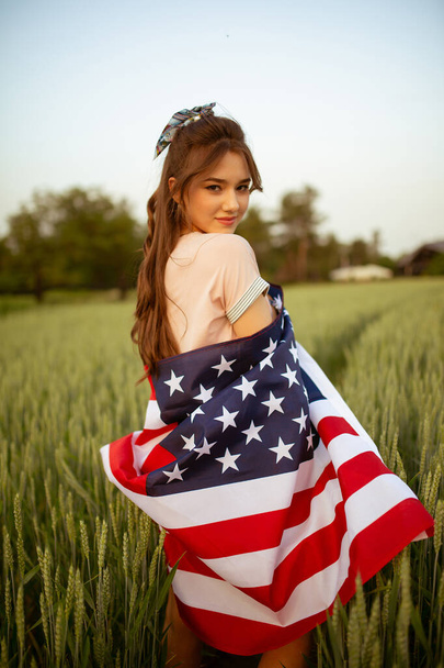giovane felice bella ragazza adolescente caucasica in top rosa e pantaloncini jeans con bandiera nazionale americana su di lei nel campo di grano verde al tramonto. Giorno di indipendenza - Foto, immagini