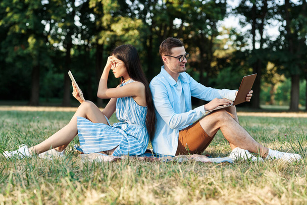 ノートパソコンとタブレットを持って芝生の上に座っている若い学生は  - 写真・画像