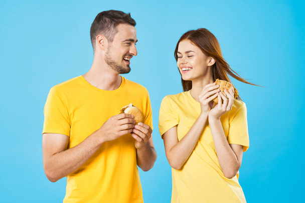 Young beautiful hungry couple eating hamburgers in studio. Isolated - Fotografie, Obrázek