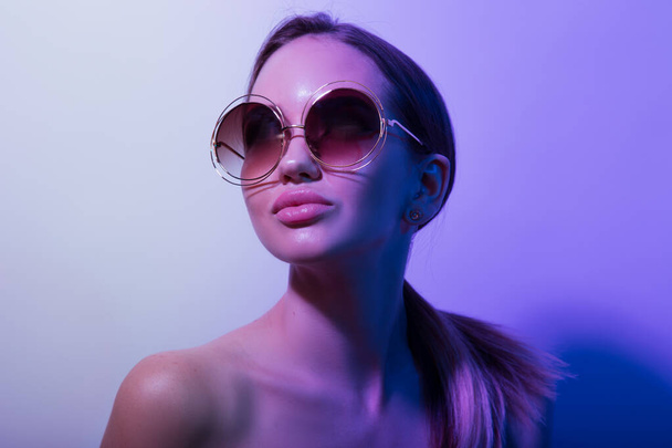 Κορίτσι της μόδας με στρογγυλά γυαλιά που ποζάρουν σε ένα νέον φως στο στούντιο. Πορτρέτο - Φωτογραφία, εικόνα