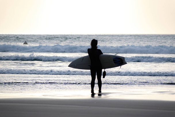 View of young woman surfer wearing black wetsuit and holding surfboard, preparing to enter Piha beach surf - Photo, Image