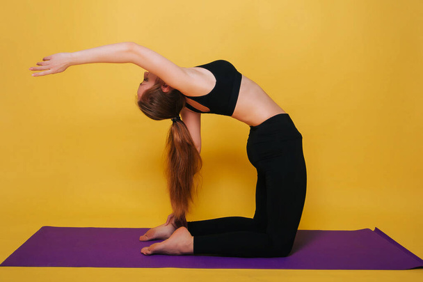 Athletische, schöne junge Mädchen beim Yoga in Uschtrasana-Pose. Tragen Sie schwarze Sportbekleidung auf einem schlichten gelben Hintergrund. - Foto, Bild