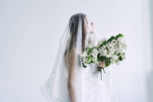Young woman under plastic sheeting holding flowers. Ecology concept - Foto, afbeelding