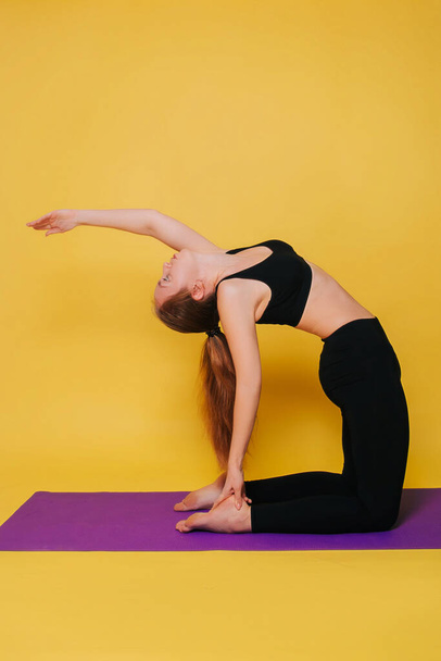 Атлетичная, красивая молодая девушка, практикующая йогу в позе уштрасаны. Носите черную спортивную одежду на просторном желтом фоне
. - Фото, изображение