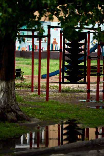 Pfütze nach Regen in der Nähe des Sportplatzes, unter einem großen Baum. Es gibt keine Menschen. Leerer Spielplatz. - Foto, Bild