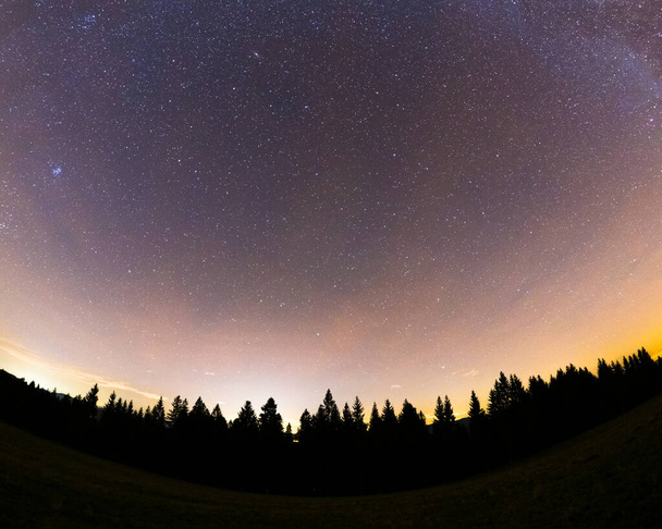 Ο Γαλαξίας μας συνέλαβε την ευρεία εξάπλωση μαζί με τις κορυφές των δέντρων με μια κίτρινη-πορτοκαλί λάμψη στον ορίζοντα και πολλά αστέρια στον ουρανό κατά τη διάρκεια μιας ολόκληρης νύχτας. - Φωτογραφία, εικόνα