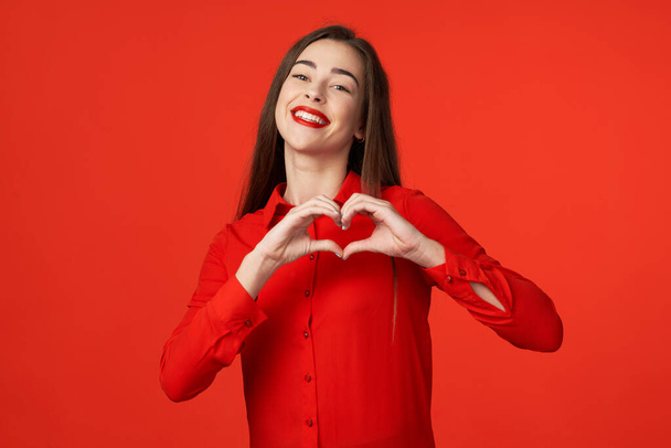 Een studio opname. Jonge mooie vrouw in rood met hart gebaar geïsoleerd op rode achtergrond - Foto, afbeelding