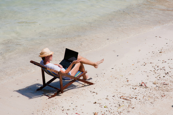 Homem sentado em uma cadeira de praia com um laptop na praia
 - Foto, Imagem