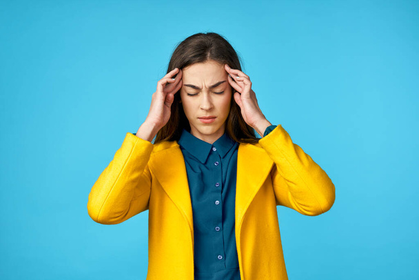 молода красива жінка в жовтій куртці з головним болем в студії з синім тлом
. - Фото, зображення