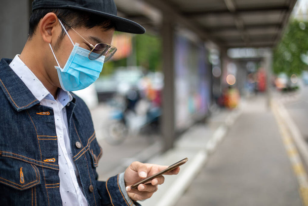 Aasialainen nuori mies, joka käyttää älypuhelinta kaupungissa ja käyttää kasvonaamiota ilman pilaantumisen, hiukkasten ja influenssaviruksen, influenssan, koronaviruksen suojaamiseen
 - Valokuva, kuva
