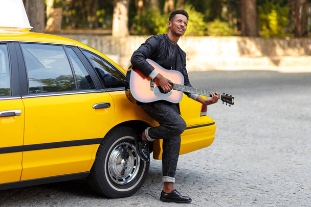 Un retrato de vista lateral de tipo de raza múltiple en ropa casual cerca de coche vintage amarillo, jugar a la guitarra, en la calle
. - Foto, imagen
