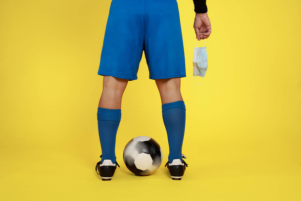 football player with face mask in hand due to covid19 coronavirus pandemic and ball between legs on yellow background - Photo, Image