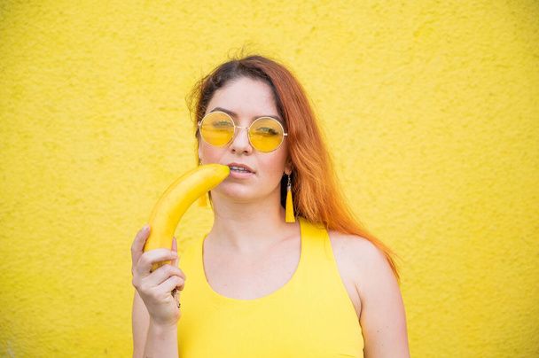 Portrait of a beautiful red-haired woman in a yellow dress and sunglasses holds a banana near sensual lips. Girl posing with fruit on a yellow background. - Fotoğraf, Görsel