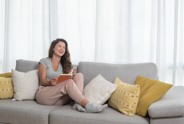 Young beautiful girl or woman lying on the sofa at home reading the book and enjoying her free time without her husband and children with smile and happy face - Zdjęcie, obraz
