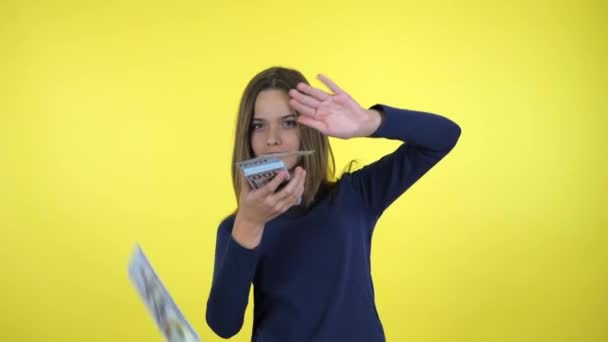 Rich teenager girl throws lot of money and smiles on yellow background - Imágenes, Vídeo