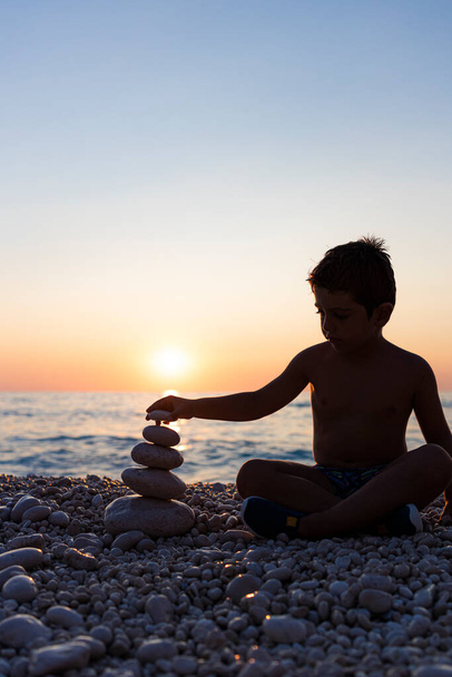 Pieni lapsi leikkii kivillä rannalla auringonlaskun aikaan
 - Valokuva, kuva