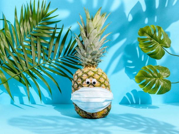 Summer 2020: Cute pineapple with eyes in medical mask among palm leaves with tropical shadows on blue background - Foto, afbeelding