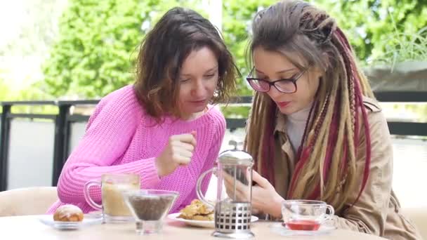 Modern meisje met lange dreadlocks toont iets op de smartphone aan haar mooie vriendin in roze trui tijdens een lunchpauze samen in een coffeeshop buiten - Video