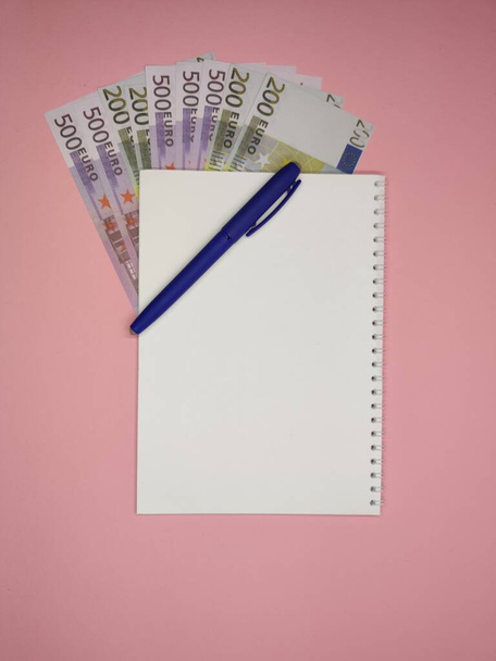 on a pink background, a blue pen on the left diagonally on a Notepad with a copy space, 200 and 500 Euro bills peek out from under it. - Φωτογραφία, εικόνα