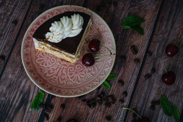 Piece of cake with cream on plate. Sweet cake on wooden background with coffee beans, cherry and mint leaves. Delicious slice of layered tart on plate. Baking and decorating cake - Foto, Imagem