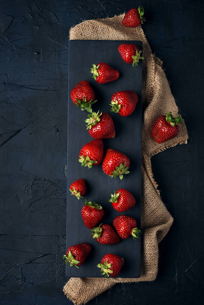 Ripe red strawberries with black serving board on burlap and dark blue background. Sweet dessert from fresh berries on the table. Top view.  - Fotó, kép