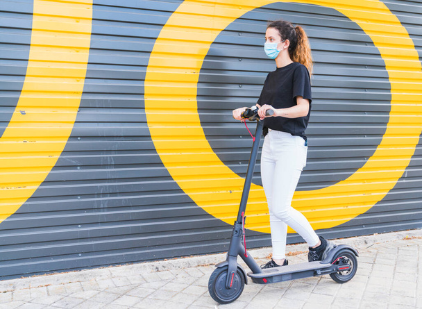 Nieuw normaal. Jong meisje met een masker met een elektrische scooter. Vrouw die gebruik maakt van elektrisch en ecologisch transport Veiligheid en pandemie concept. Coronavirusapparaten. Menselijk leven op sociale afstand - Foto, afbeelding