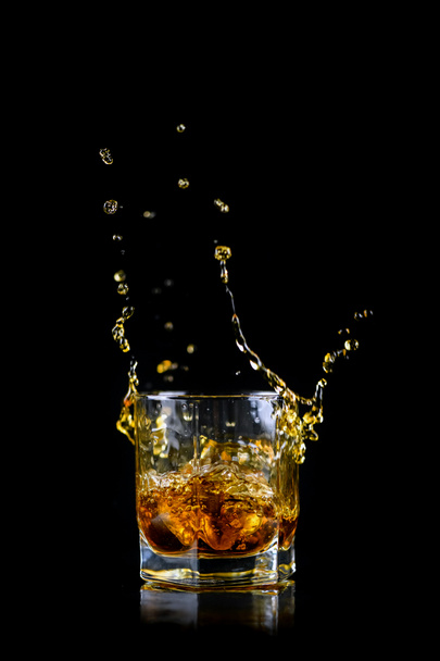 Glass of splashing whiskey or other alcohol with ice cube isolated on black background - Foto, afbeelding