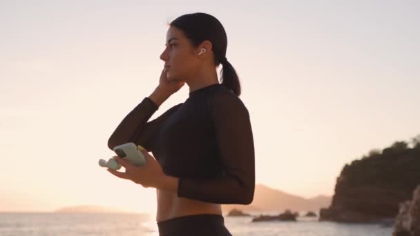 sportowa kobieta słucha muzyki na plaży o zachodzie słońca - Materiał filmowy, wideo