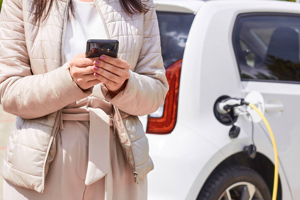 Woman with a mobile phone near recharging electric car. Vehicle charging at public charging station outdoors. Car sharing concept. - Photo, Image