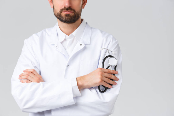 Лікарня, медичні працівники, концепція лікування ковадла-19. Обрізаний знімок серйозно впевненого лікаря з бородою, одягнений в білі скраби. Лікар хрест руки груди, тримає стетоскоп
 - Фото, зображення