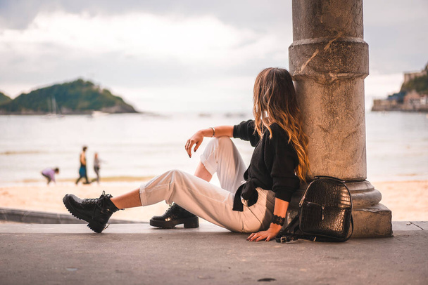 Mode de vie en ville avec une fille blonde en pantalon blanc et pull en laine noire près de la plage. Assis sur les hauts-fonds d'une plage sur un beau coucher de soleil
 - Photo, image