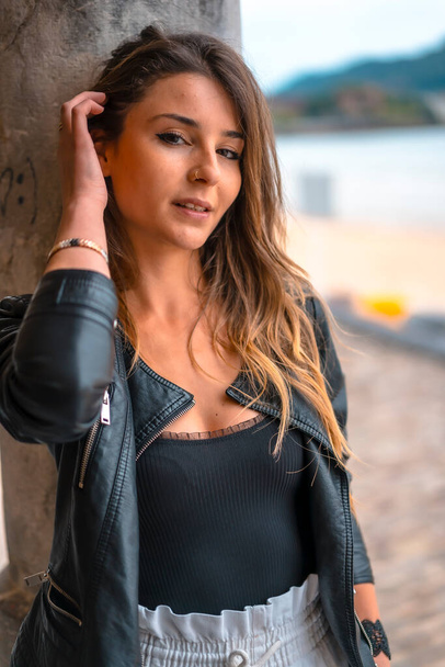 Lifestyle in the city with a blonde girl in white pants and a leather jacket near the beach. Close-up portrait putting on hair - Foto, afbeelding