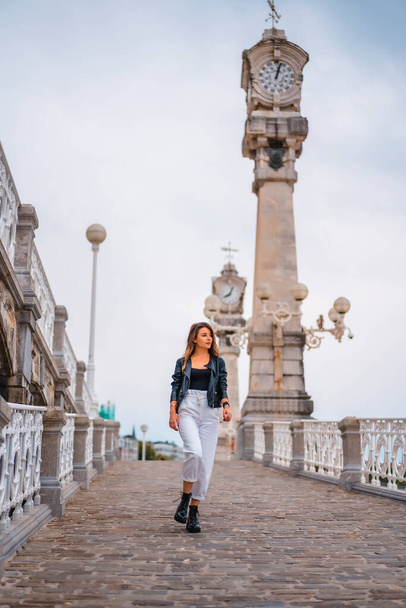 Lifestyle in the city with a blonde girl in white pants and a leather jacket. Walking down a slope with beautiful clocks in the background - Фото, изображение