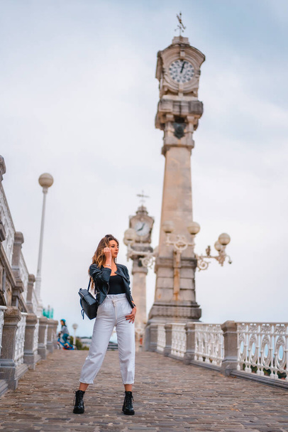 Lifestyle in the city with a blonde girl in white pants and a leather jacket. Perched on a hill with clocks in the background near the beach - Φωτογραφία, εικόνα