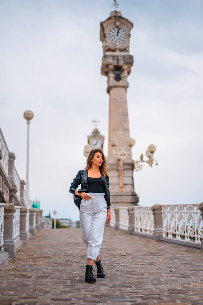 Lifestyle in the city with a blonde girl in white pants and a leather jacket. Walking down a slope with clocks in the background near the beach - Фото, изображение